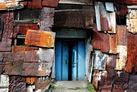 In the center are blue doors that are surrounded by orange, white, and brown pieces of metal-looking material. This is what a warehouse looks like in a Mumbai slum.