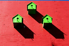 A photo of three green birdhouses casting shadows on a red wall, illustrating an article about exclusionary practices in housing