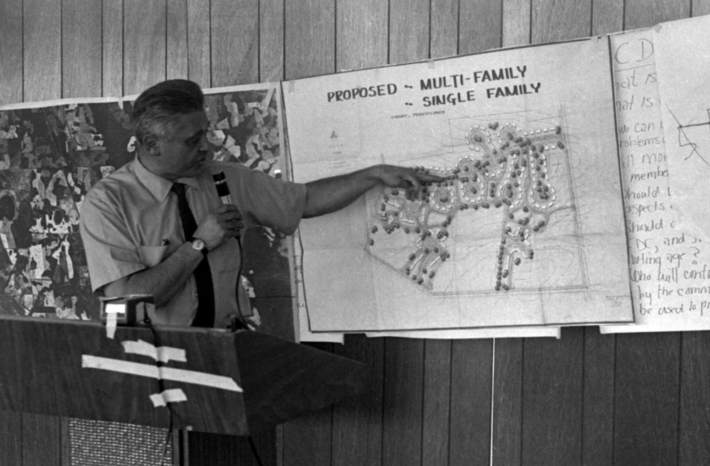 A black and white photo of a man pointing to a design of a multi-family residences called New Communities.