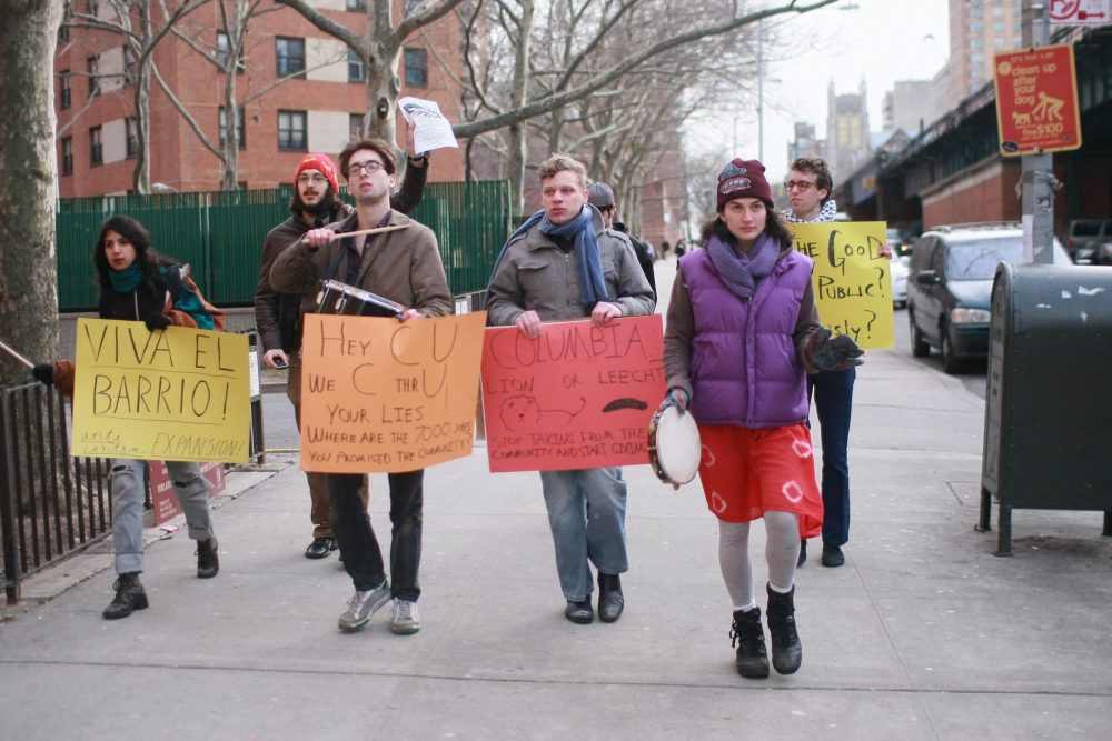 College students walk down a street in Harlem holding signs that show their displeasure with Columbia University. Some students are playing musical instruments.