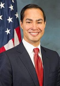 A man, who is wearing a red tie, white shirt, and a dark colored jacket, smiles. In the background, there's a partial view of the American flag. Less than 10 stars and partial red and white strips can be seen.