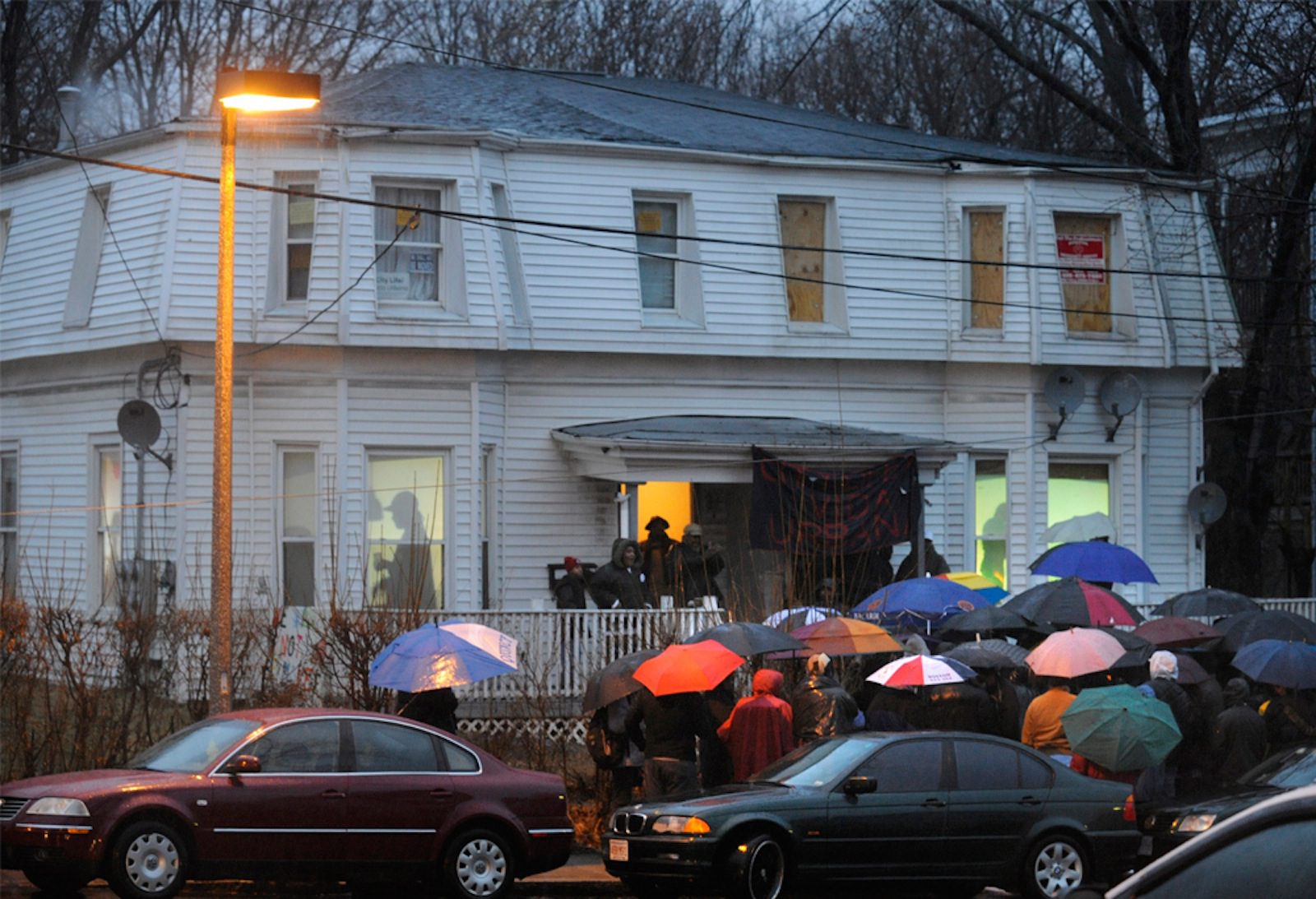 People stand outside of a home in the rain while a projection of people are shown inside. This was part of an art installation in Boston.