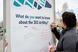 A woman writes a question on a whiteboard that asks "What do you want to know about the 303 ArtWay?
