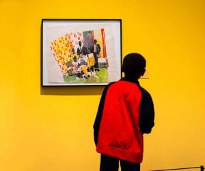 A young boy wearing a red and black jacket looks at a painting of a group of people, which is placed in a room that has bright yellow walls.