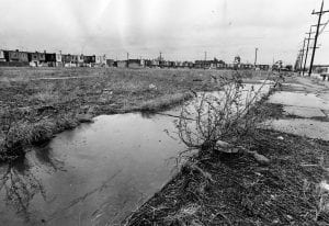 An empty lot, bordered by houses on the left and utility poles on the right. In the center is a ditch which has accumulated water. The site is a lot on which the Whitman Park Houses were built.